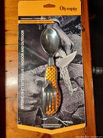 Front view of Olympia multi cutlery in blister packaging showing spoon, fork, and knife functions with orange handle.