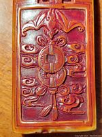 Close-up of the rectangular cultured wood Buddhist amulet featuring bat and coin motifs in concave/convex design.