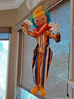 Front view of the vintage Pelham Puppets clown marionette puppet hanging by its strings. Shows fabric costume with orange, blue, white, brown vertical stripes, ruffled collar, and painted face with blue hair.