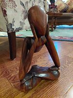 Left side angle view of vintage wood carved abstract figure seated in Padmasana meditation pose on wooden floor, with upholstered furniture in background.