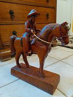 Front angled view of vintage wood carved cowboy on horse sculpture with leather saddle, showing detailed carving and polished finish.