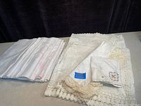 Pair of short crocheted lace curtains, crocheted doilies, and two embroidered cloths with crochet edges all laid out to show details and condition.