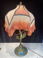 Full view of the lamp showing pink and orange fabric shade with thick fringe trim and bronze lady figurine base.