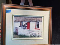 Framed photograph showing a horse standing in front of a white thatched-roof cottage with red door and window trim behind a stone wall, framed in wooden frame with beige matting.