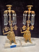 Full view of the pair of gilded brass antique candle holders with Czechoslovakia crystal prisms on white marble bases against black background.