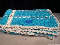 Folded turquoise and cream crocheted throw blanket on a grey surface with a black background. Showing texture and handmade crochet pattern of the blanket.