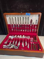 Open wood box with red velvet and white satin lining showing the full cutlery set. Knives, forks, spoons, some with decorative handles laid in place.