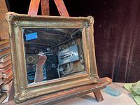 The photo shows a rectangular wall mirror in an antique gilded frame with decorative corners and visible age wear. It is resting on a wooden easel and reflects a background featuring tools and a fuse box.
