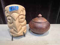 Front view of the carved wood mask and antique wood bowl with lid closed, showing the mask's stylized facial features and bowl's rounded form.