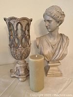 Photo showing both bust of woman and ornate vase-like candle holder with taupe pillar candle in front