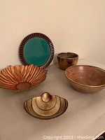 Five decorative bowls and dishes arranged on a surface: green and brown plate, wooden bowl, copper-colored planter, gold flower-shaped glass bowl, shell-shaped dish.