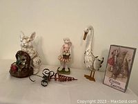 Group photo showing all Easter decor items together on a white surface against a plain wall. Items include porcelain bunny with basket, girl bunny figurine, metal goose, metal carrot, and Easter greeting card.