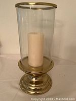 Photo of a large brass candle holder with a tall clear glass chimney and white pillar candle inside. The holder has a stepped base design with reflective brass finish.