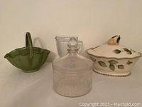 Photo shows all four items: green ceramic basket with handle, a clear etched glass jar with stopper, clear textured water jug, and a cream ceramic covered serving bowl decorated with leaves and a pear-shaped handle on the lid.