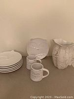 Photo of six stacked white ceramic dessert plates, two mugs, one water jug against neutral background, showing owl motif details.