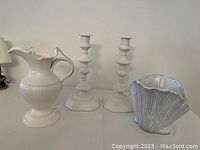 Photo showing the complete set of a white porcelain pitcher with ornate handle, two matching candle holders, and a shell-shaped bowl all arranged on a flat surface against a white wall.