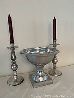 Front view of two silver plate candlesticks holding dark red candles and a silver plate pedestal bowl with square base, all arranged against a plain wall.