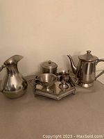 Full set of six silver plate serving items on display against a plain wall including hexagonal tray, water jug, teapot, creamer, sugar bowl with lid, and a small lidded pot.