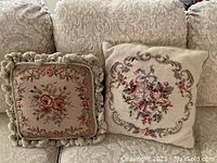 Two tapestry design square throw pillows on a beige patterned couch: one 12-inch pillow with fringe and floral design, one 15-inch needlepoint floral pillow