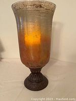 Front view of bronze/gold glass vase-shaped candle holder with crackled texture and brown ornate base, lit with battery candle inside.