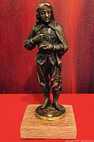 Front view of the bronze figurine standing on a round base against a red background, showing detailed facial features and clothing.
