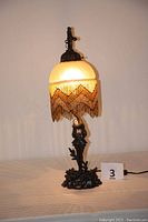 Photo of illuminated lamp showing the beaded fringe shade and figurine base on a light background.