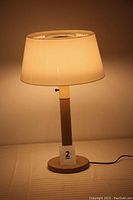 Full view of illuminated mid-century modern table lamp with cylindrical metal base and light-colored plastic shade emitting warm light, placed on table.