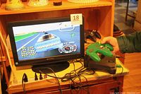 Photo of the Nintendo 64 console powered on with NASCAR 99 gameplay displayed on the Sansui 19" TV, with one green controller held by a hand and connected to the console.