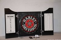 Front view of the open dart board cabinet showing the Harley-Davidson dart board in the center and score sheets on each side, with six darts and mounting screws on the table beneath.