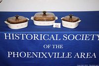 Set of three CorningWare casserole dishes with Pink Hearts Forever Yours pattern and brown tinted glass lids on a blue tablecloth with Historical Society text.