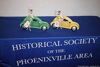 Pair of two handmade jewelry organizers displayed on blue cloth with Historical Society banner. Each features a girl with wire loops for hanging earrings, bracelets, and necklaces, standing beside vintage cars in green and yellow.