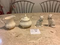 Full view of Royal Albert creamer, sugar bowl with lid, Beleek cat figurine, and a second non-branded cat figurine placed on a marble tabletop