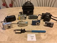 Full lot showing all 8 cameras, black Icon camera bag, and blue-handled selfie stick on table.