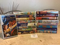 Photo showing 16 VHS tapes stacked in two rows, titles visible include Beauty and the Beast Special Edition, The Lion King, The Wizard of Oz, The Land Before Time VIII, Alice in Wonderland, A Charlie Brown Christmas, Rudolph and the Island of Misfit Toys, Aladdin, The Little Rascals, Barbie Rapunzel, Pippi, Snow White and the Seven Dwarfs, Rocket Power.