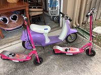 Purple vintage-style electric scooter alongside two pink Razor kick scooters on concrete floor outside garage.