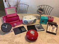 Photo of all lot items arranged on a table including CD player, clock, calculators, cosmetic organizer, nail dryers, desk bell, pencil sharpener, vanity mirror, and bottles.