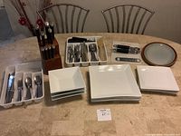 Photo showing white square dinner plates and bowls stacked, stainless steel utensils organized in a plastic organizer, a wooden knife block with knives, and various kitchen tools on a countertop.