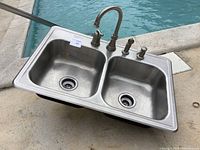 Front view of stainless steel dual basin sink with Moen faucet attachments mounted on deck, showing the basin interiors and drain holes.