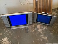 Two flat-screen TVs side by side on floor with remotes visible under them, showing screen fronts and bases.