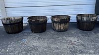 Front view of 4 rustic round wooden planters with black metal bands, aligned side by side on the ground, showing weathered surfaces and rustic outdoor wear.