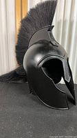 Side view of black metal Achilles helmet with plume crest on black surface and corrugated background