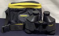 Black Bushnell carrying case with yellow zipper and Bushnell logo patch, alongside a pair of black binoculars with attached shoulder strap.