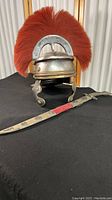 Front angled view showing Roman Centurion helmet with red plume and decorative sword laid on black surface.