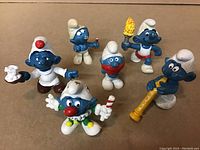 Photo showing six vintage 1980s Smurfs figures including various poses and accessories, placed on a brown surface.