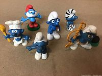 Photo showing six vintage Smurfs figurines on a beige surface, including a chef, shepherd, zebra-striped, seated and other character Smurfs