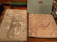 Photo showing two sets of packaged Italian cotton tablecloth and napkin sets, folded inside original boxes with visible floral embroidery on tablecloth edges.