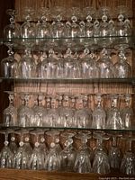 Shelves filled with multiple rows of clear glass stemware including wine glasses and goblets showing good condition with some dust.