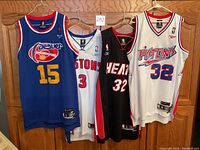 Four stitched NBA jerseys on hangers against a wooden background, showing front views.
