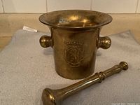 Side view of solid brass mortar and pestle, showing the entire mortar with two side handles and the pestle resting beside it on a grey cloth surface.