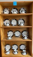 Full shelving unit showing four rows of Mikasa Charisma Black china sets including cups, saucers, and soup bowls arranged neatly.